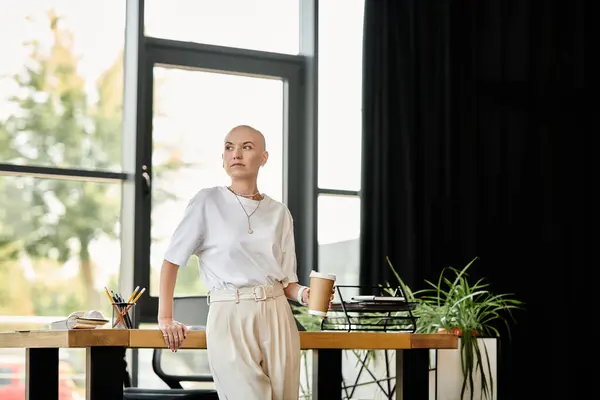 A fashionable bald woman stands confidently by the desk, savoring her drink in a chic environment. — Stock Photo