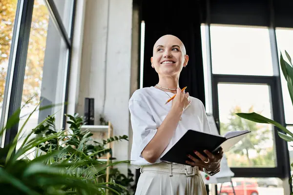A joyful young woman in stylish attire stands among greenery, holding a notebook and smiling. — Stock Photo