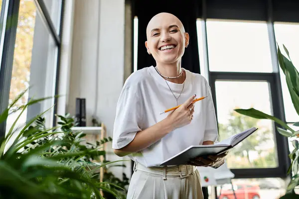 A young bald woman smiles brightly while holding a notebook and a pencil, surrounded by greenery. — Stock Photo
