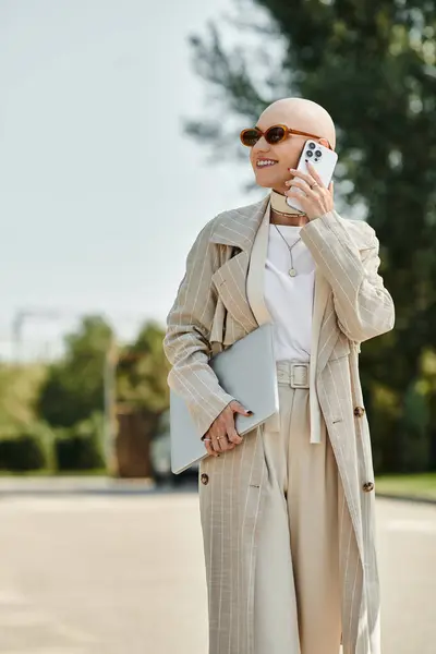 Una mujer joven y elegante con una cabeza calva sostiene un teléfono inteligente y un ordenador portátil mientras camina con gracia. — Stock Photo