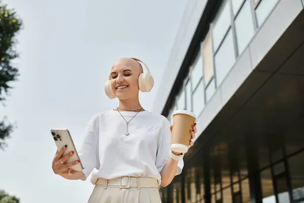 Una giovane donna calva elegante sorride gioiosamente mentre si gode caffè e musica all'aperto. — Stock Photo