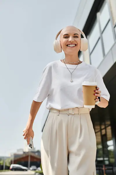 A young bald woman dressed elegantly walks joyfully outdoors, wearing headphones and holding coffee. — Stock Photo