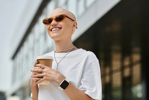 A young woman with a bald head smiles brightly while enjoying coffee in stylish attire outside. — Stock Photo