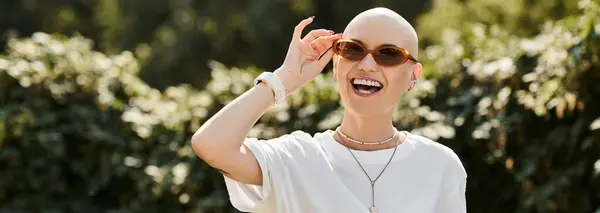 A young bald woman in elegant attire playfully adjusts her sunglasses while smiling outdoors. — Stock Photo