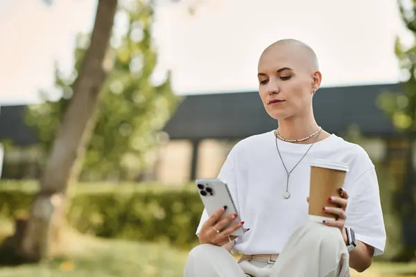 A bald woman dressed elegantly relaxes outdoors, sipping coffee and checking her phone. — Stock Photo
