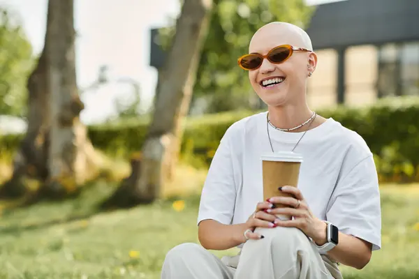 A cheerful bald woman in stylish attire relaxes outdoors, savoring her coffee amidst greenery. — Stock Photo
