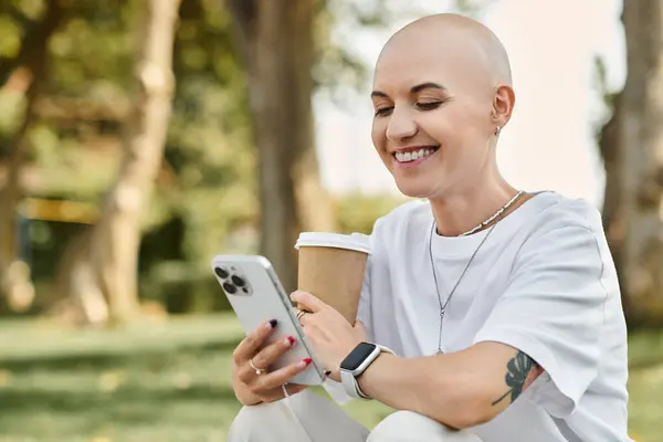 A cheerful bald woman dressed elegantly sips coffee while engaging with her phone outdoors. — Stock Photo