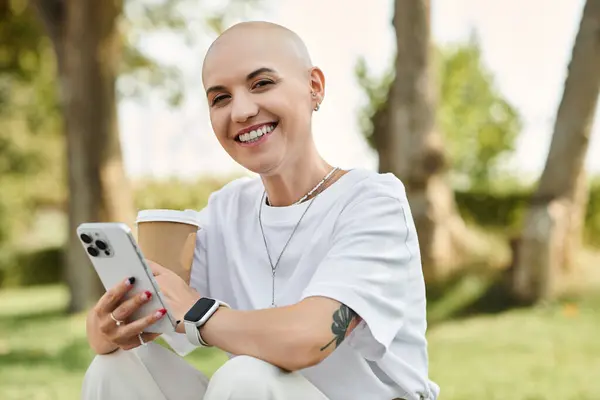 A young bald woman dressed elegantly sits in a park, smiling and texting on her phone. — Stock Photo