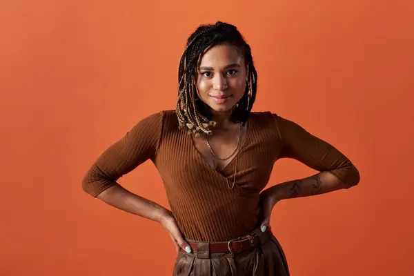 A young African American woman with braided hair stands confidently against a bright orange backdrop, exuding style. — Stock Photo