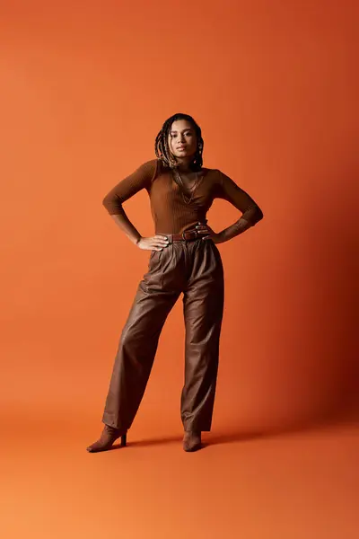 A young woman with braids stands confidently in a studio, showcasing her fashionable outfit against a warm backdrop. — Stock Photo