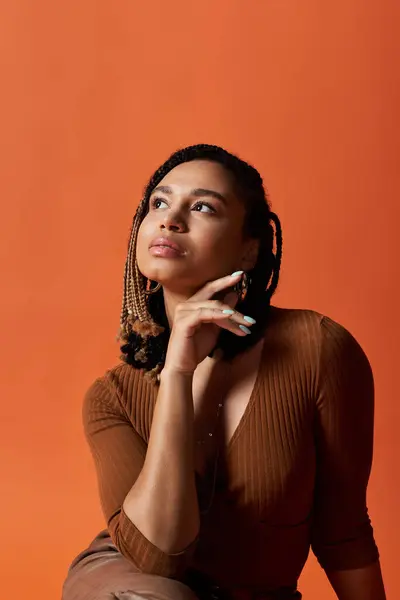 A fashionable young woman with braided hair thoughtfully poses against a bold, warm backdrop in a studio. — Stock Photo