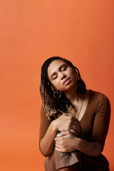 Una giovane donna afroamericana con trecce alla moda posa elegantemente su un audace sfondo arancione, trasudando fiducia. — Foto stock