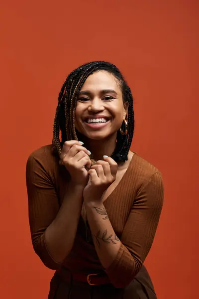 A joyful young woman with braided hair poses confidently against a striking orange backdrop, embracing the moment. — Stock Photo