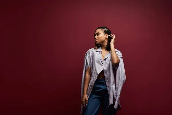 A stylish young woman with braids confidently poses in a chic studio with a rich maroon backdrop. — Stock Photo