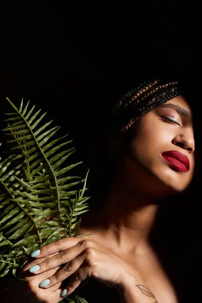 Una giovane donna con le trecce e le labbra rosse audaci posa con grazia, tenendo una felce in un ambiente dinamico studio. — Foto stock