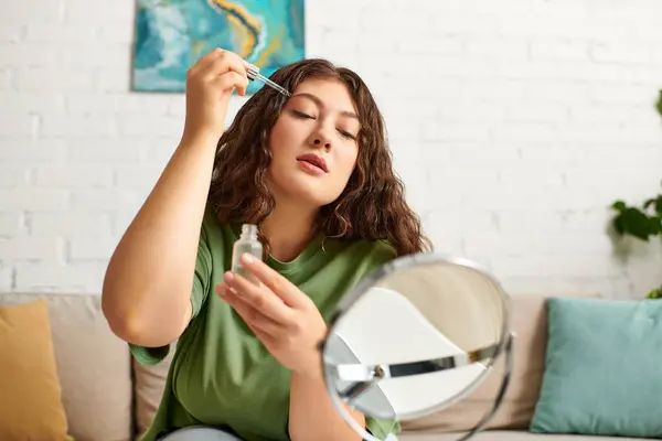 A young plus size woman with curly hair applies serum in her living room, embracing her beauty routine. — Stock Photo