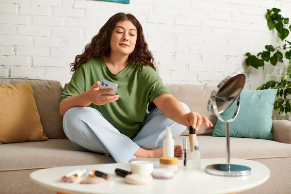 A curly plus size woman relaxes while applying makeup, surrounded by an array of beauty products at home. — Stock Photo