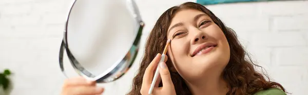 Une jeune femme aux cheveux bouclés sourit en appliquant du maquillage, immergée dans sa routine beauté décontractée à la maison. — Photo de stock