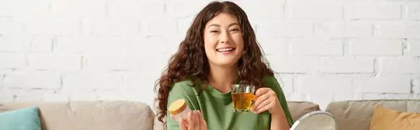 A joyful young woman with curly hair relaxes in her living room, savoring tea and beauty products. — Stock Photo