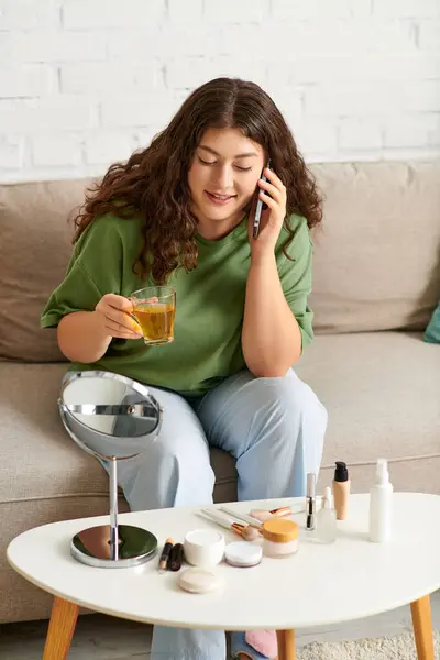 Une femme aux cheveux bouclés se détend en tenue confortable, sirotant du thé et explorant ses produits de beauté tout en parlant. — Photo de stock