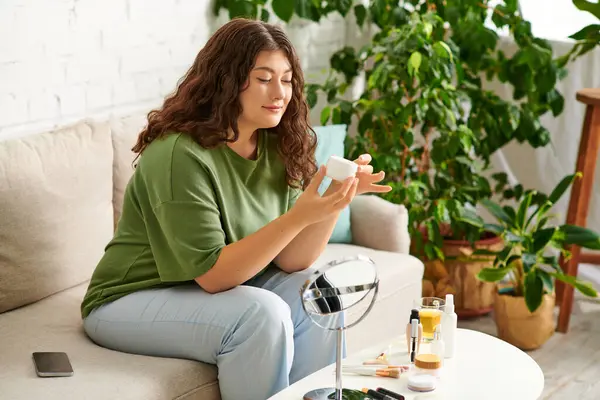 Curly plus size woman enjoys her beauty routine, applying products and relaxing in a cozy living room setting. — Stock Photo