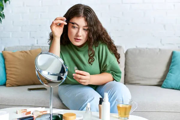 Lockige Frau in lässiger Kleidung erkundet ihre Beauty-Routine inmitten verschiedener Kosmetika in ihrem gemütlichen Wohnbereich. — Stockfoto