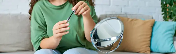 Una joven mujer de talla grande con el pelo rizado se relaja en su sala de estar mientras se aplica maquillaje con un espejo. — Stock Photo