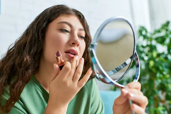 A young, plus-size woman applies lipstick while seated in her living room surrounded by beauty essentials. — Stock Photo