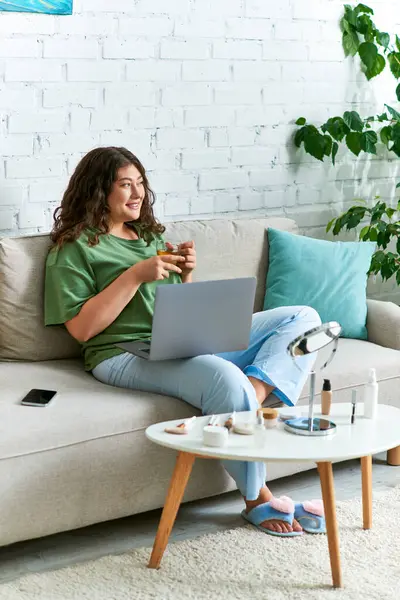 A curly plus-size woman relaxes on the couch, engaging in her beauty routine with makeup products and laptop — Stock Photo