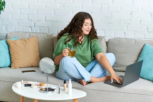 Une jeune femme aux cheveux bouclés se détend sur le canapé, s'engageant dans sa routine beauté avec ordinateur portable et thé. — Photo de stock