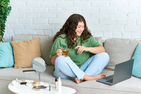 Una giovane donna plus-size si rilassa in un abito casual, godendo la sua routine di bellezza con prodotti per la cura della pelle e laptop — Foto stock