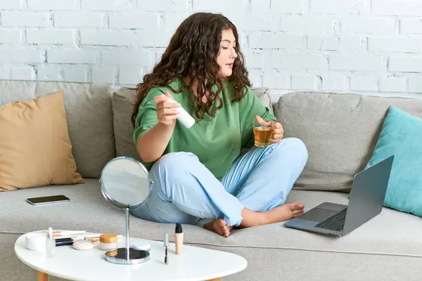 Une jeune femme frisée se détend dans son salon, s'engageant dans sa routine beauté tout en dégustant une boisson chaude. — Photo de stock