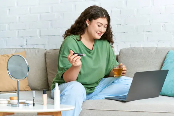 Une jeune femme de taille plus aux cheveux bouclés se détend dans son salon, sirotant du thé et essayant des cosmétiques. — Photo de stock