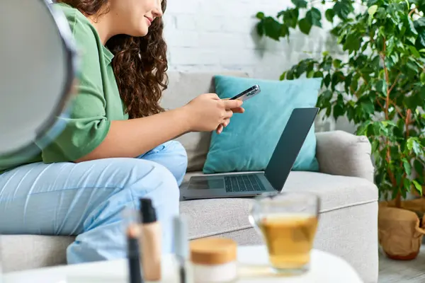 Une jeune femme aux cheveux bouclés se détend dans son salon, engagée dans sa routine beauté entourée de cosmétiques et d'appareils — Photo de stock