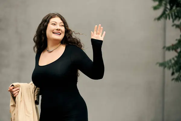 Brightly smiling, a young woman in a black dress waves warmly while enjoying the autumn air outdoors. — Stock Photo