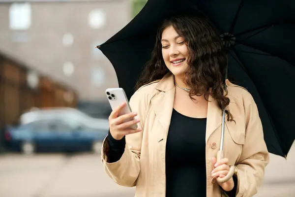 A young woman in a black dress and beige trench coat smiles as she checks her phone while standing outside. — Stock Photo
