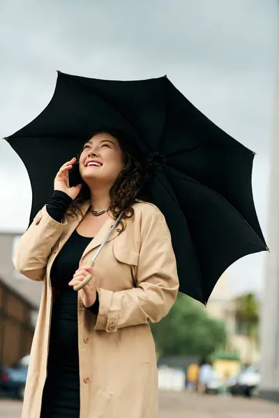 A young plus-size woman in a black dress and trench coat smiles while talking on the phone amid autumn vibes. — Stock Photo