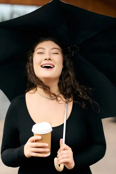 A young woman strolls outdoors on an autumn day, sipping coffee while holding an umbrella and smiling beautifully. — Stock Photo