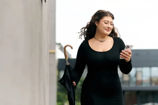 A young woman in a black dress strolls outdoors, checking her phone on a cool autumn afternoon. — Stock Photo