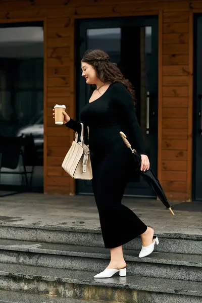 A young woman strolls outdoors in a stylish black dress, enjoying an autumn day with a warm drink in hand. — Stock Photo