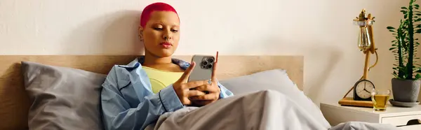 A young woman sits comfortably in bed, scrolling through her smartphone after a busy day. — Stock Photo