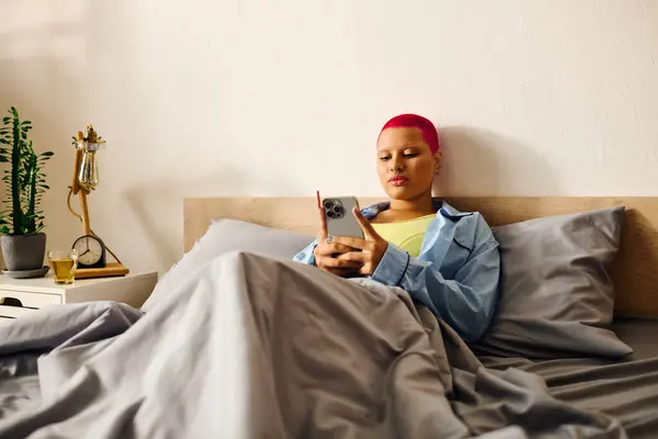 A young woman sits comfortably in bed, engaged with her smartphone while wrapped in blankets. — Stock Photo