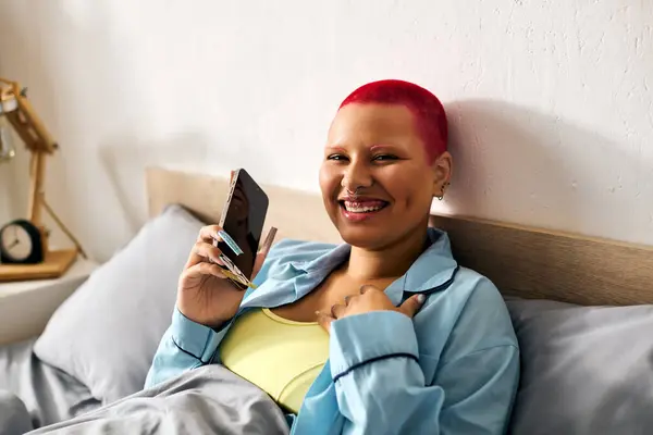 A young woman relaxes in bed, happily engaging with her phone and sharing a joyful smile. — Stock Photo