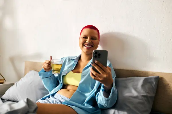 Uma jovem se deita em sua cama, sorrindo enquanto segura uma bebida e verifica seu telefone, irradiando alegria. — Fotografia de Stock