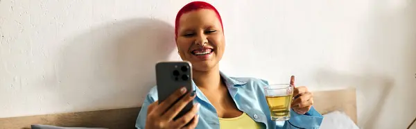 A young woman relaxes at home, sipping a drink, and smiling at her phone while unwinding. — Stock Photo