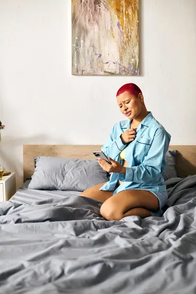 A young woman sits on her bed, expressing deep feelings while holding her phone in a cozy space. — Stock Photo