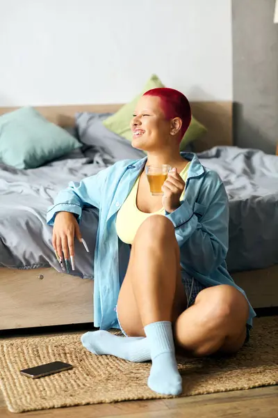 Una joven alegre se sienta casualmente en el suelo, saboreando su tiempo en casa y disfrutando de un capricho. - foto de stock