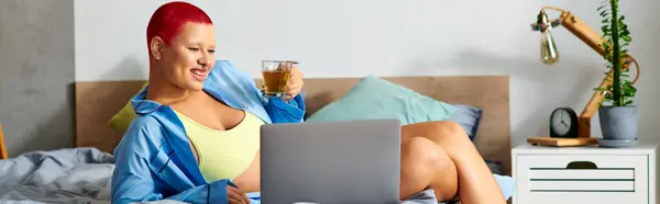 A young woman with vibrant red hair comfortably sits in bed, sipping a drink while using her laptop. — Stock Photo