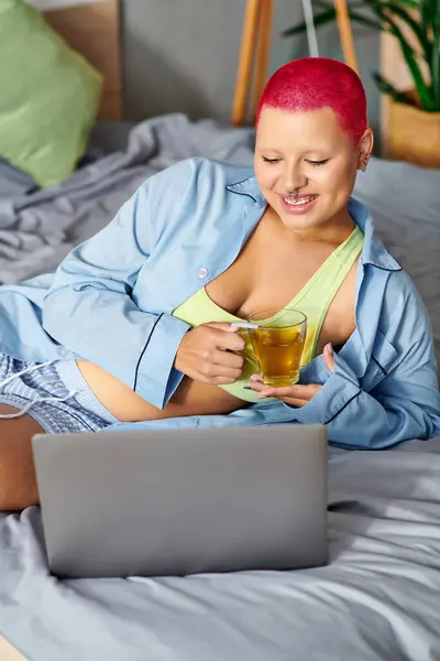 Sorrindo jovem mulher lounges em sua cama, tomando chá e usando seu laptop em uma atmosfera acolhedora. — Fotografia de Stock