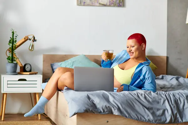 In a cozy bedroom, a young woman sips tea, lounging on her bed with a laptop open. — Stock Photo